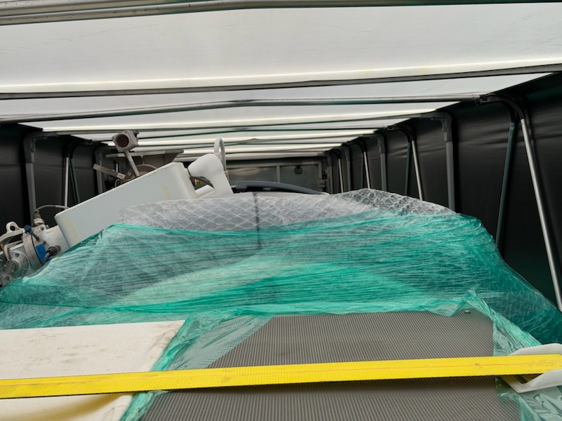 Interior of a Conestoga trailer showing roof bows, curtain rails and a yellow strap over shrink-wrapped machinery