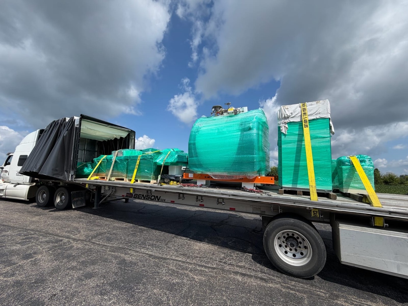Conestoga trailer rolled back showing a shrink-wrapped process tank on a skid and palletized crated equipment strapped to the deck