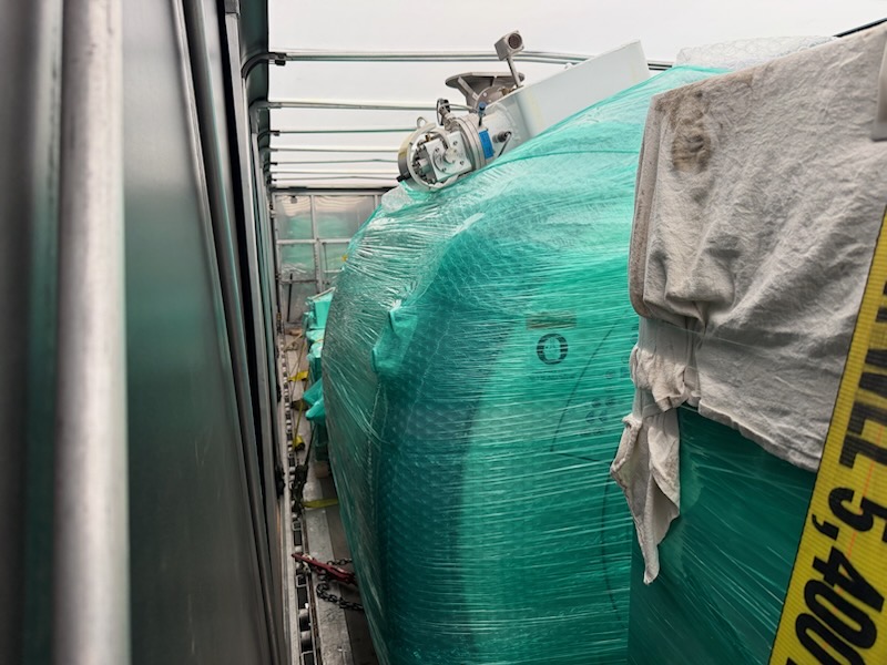 Shrink-wrapped industrial machinery secured inside a Conestoga trailer with a 5,400 lb working load limit strap and chain tie-downs