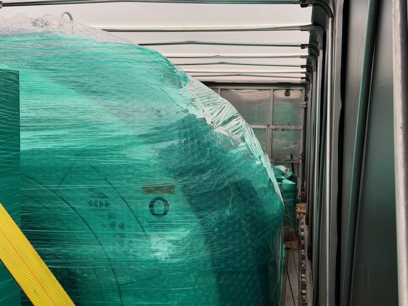 Green shrink-wrapped equipment loaded inside a Conestoga curtain-side trailer, viewed down the deck