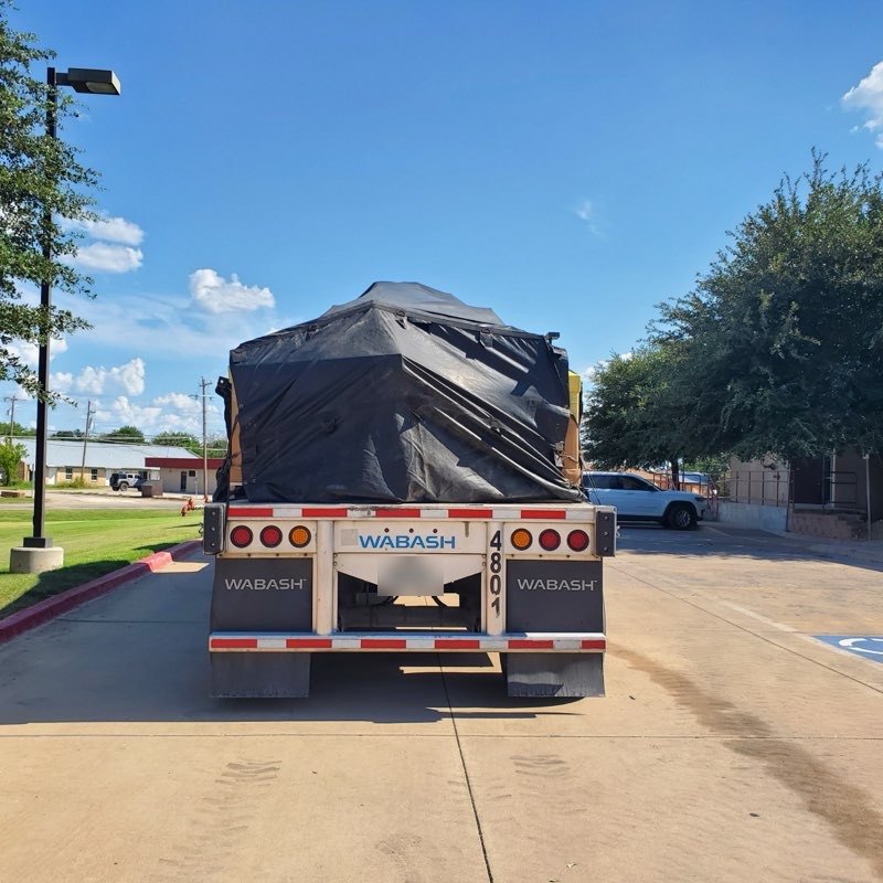 Rear view of a Wabash flatbed trailer with machinery tarped in black and strapped down for over-the-road transport